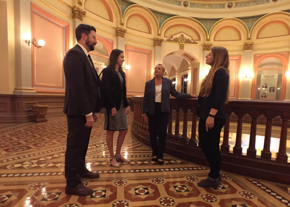 Four adults, three women and one man, dressed in professional attire standing together and facing each other by a balcony railing inside the California State Capitol building.